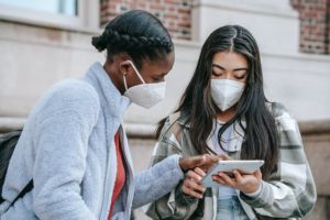 Two young women wearing face masks share a tablet. 